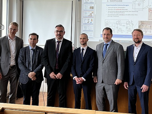 In a seminar room in front of a projector screen with a presentation, the participants in the doctoral defense stand in a row for a group picture.