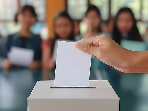 Young people are sitting in the lecture hall in the background, in front of them you can see a hand inserting a white piece of paper into a ballot box.