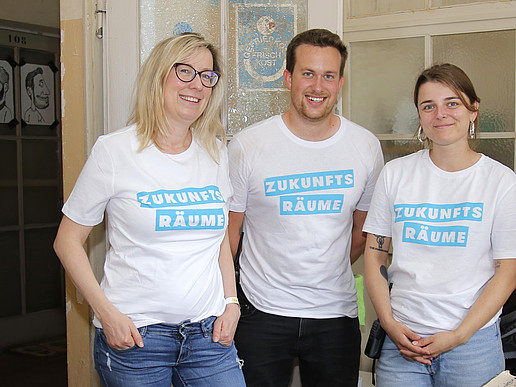 Drei Personen mit einheitlichem T-Shirt mit der Aufschrift Zukunftsräume stehen in einem Duftseminarraum.