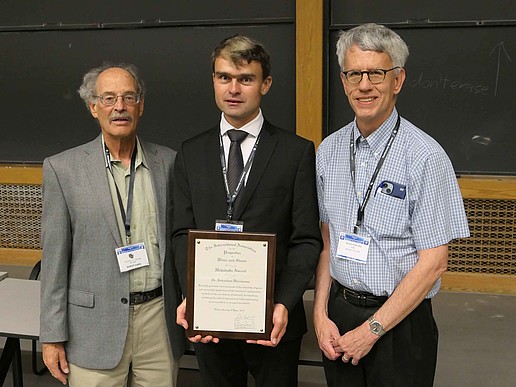 Three men stand next to each other. Dr. Herrmann in the middle holds a certificate in his hands.