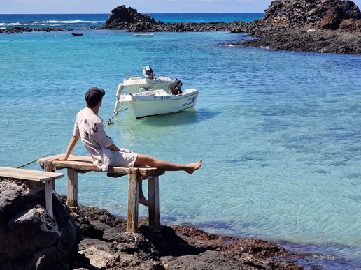 A young man sits on a bench on the beach. A white boat floats in the bay on the azure blue water in front of him.