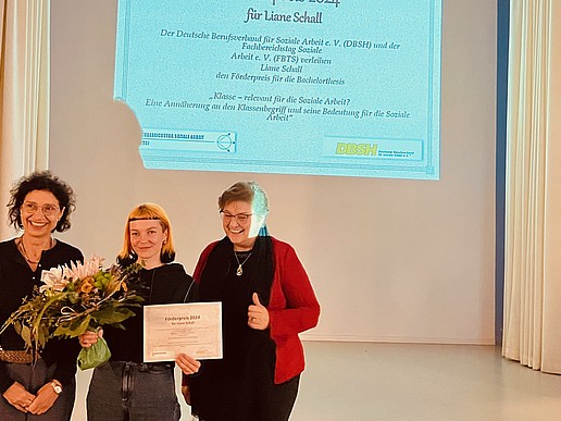 Three women stand in front of a projector screen. In the middle is the award winner with her certificate.