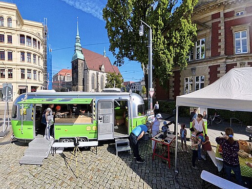Der Mobile Lernort-Anhänger steht mit offenen Türen und Seitenfenster auf dem Görlitzer Postplatz. Daneben steht ein weißer Pavillon. Darunter beschäftigen sich Kinder und andere Gäste mit den ausgelegten Materialien.