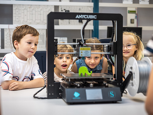 Four children are sitting in the electrical engineering laboratory looking at a small green elephant on the platform of a device.