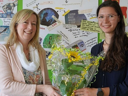 The Vice-Rector and Tanja Schwarz hold a bouquet of flowers between their hands in front of a pinboard.