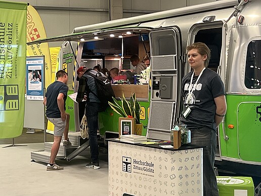 Der Airstream-Anhänger steht in der Messehalle und Besuchende betreten ihn und schauen sich Experimente an. Im Vordergrund steht eine Mitarbeiterin an einer HSZG-Theke.