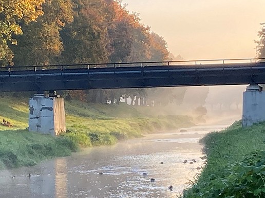 A bridge leads over a river with autumnal trees in the background.