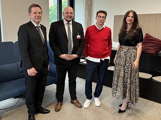 Four people stand in a room in front of chairs and look into the camera.