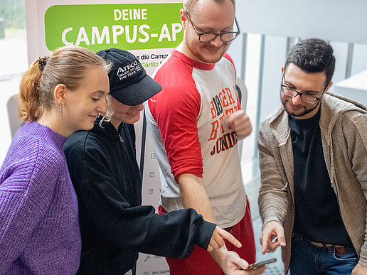 Students look at a smartphone together, in the background is a display with the caption "Your campus app".
