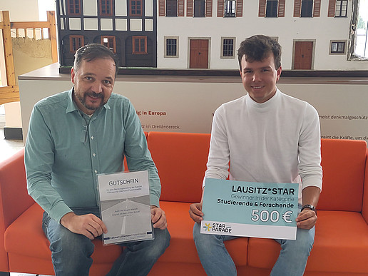 David Sauer and a student sit on a couch and hold a certificate and a check in front of them. In the background is a model of a half-timbered house.