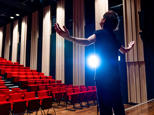 A young man stands on a theater stage, facing empty rows of red-upholstered seats.